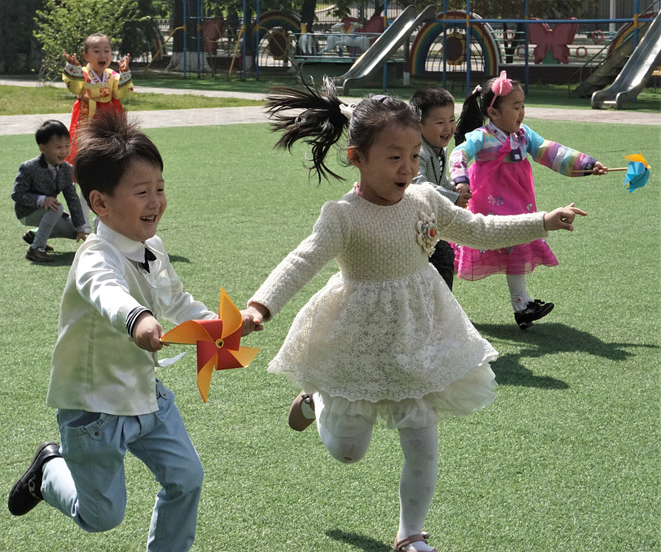 유치원 운동장에서 남녀 아이들이 짝을 지어 손을 잡고 바람개비를 돌리며 달리기 시합을 하고 있다.